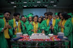 high school graduates wearing green and gold cap and gowns surround cake